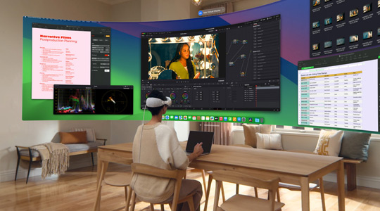 Une personne portant l’Apple Vision Pro est assise à un bureau avec un MacBook Pro 13 pouces ouvert ; un affichage incurvé et ultra grand‑angle de son écran est projeté dans la pièce