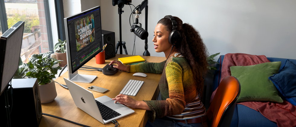 A woman in her home working at her desk with MacBook Pro connected to two external displays, wearing headphones and speaking into a microphone