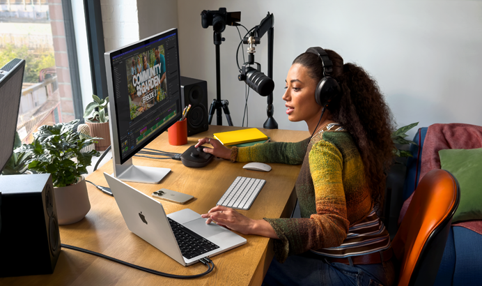 A woman in her home working at her desk with MacBook Pro connected to two external displays, wearing headphones and speaking into a microphone