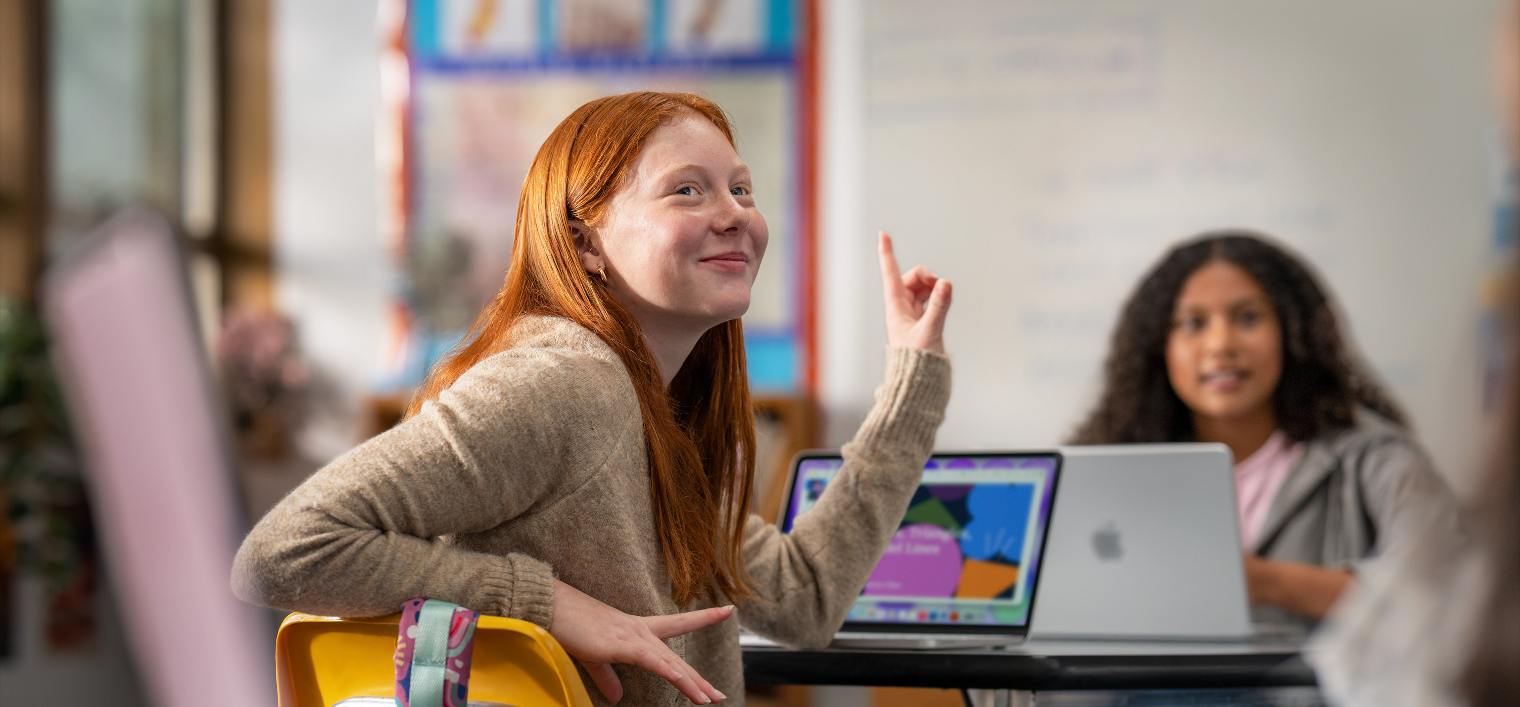 A student sits in front of a MacBook in a classroom, raising their hand to answer a question, suggesting ease of use and engagement