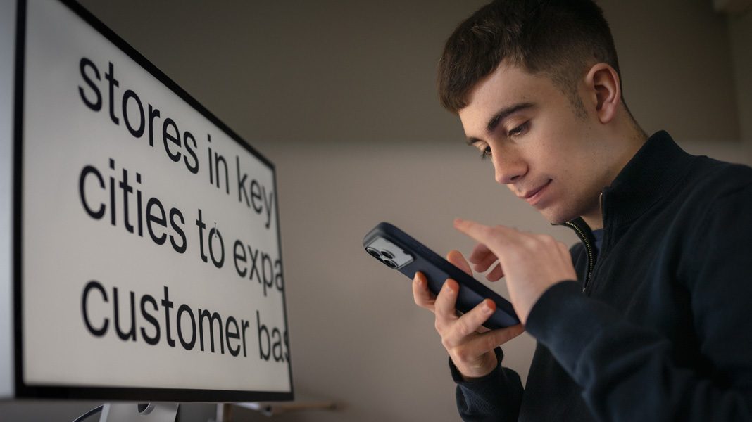 A student uses an iPhone while sitting in front of a monitor displaying content from his business class in large, zoomed-in text