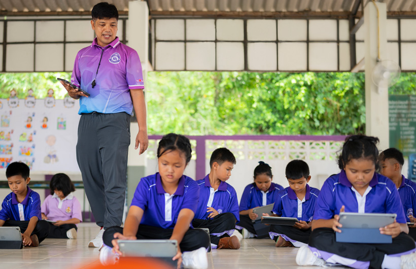 In an outdoor classroom, a physical education teacher walks among his students, who are seated on the ground using&nbsp;iPads.