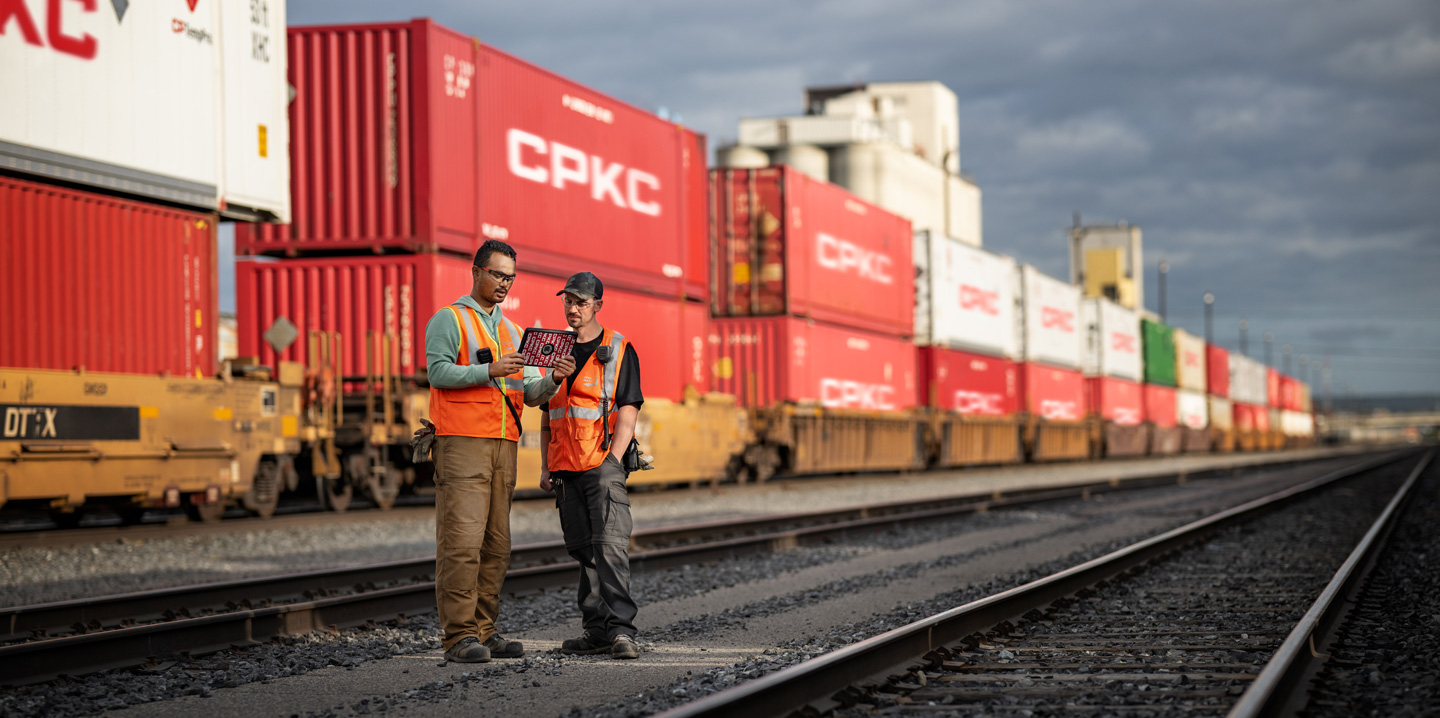 Dois trabalhadores ferroviários a olhar para um iPad no terminal ferroviário junto a um comboio parado que transporta contentores
