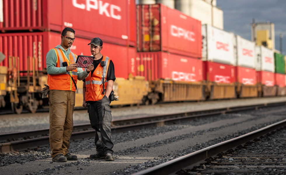 Two railroad workers reference an iPad while standing in a train depot beside a stationary train carrying shipping containers