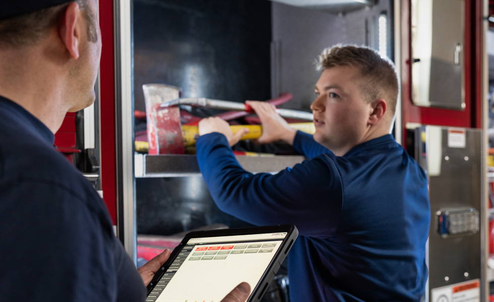Two crew members inspect tools and equipment on a fire truck, one holds an iPad while the other inspects 
