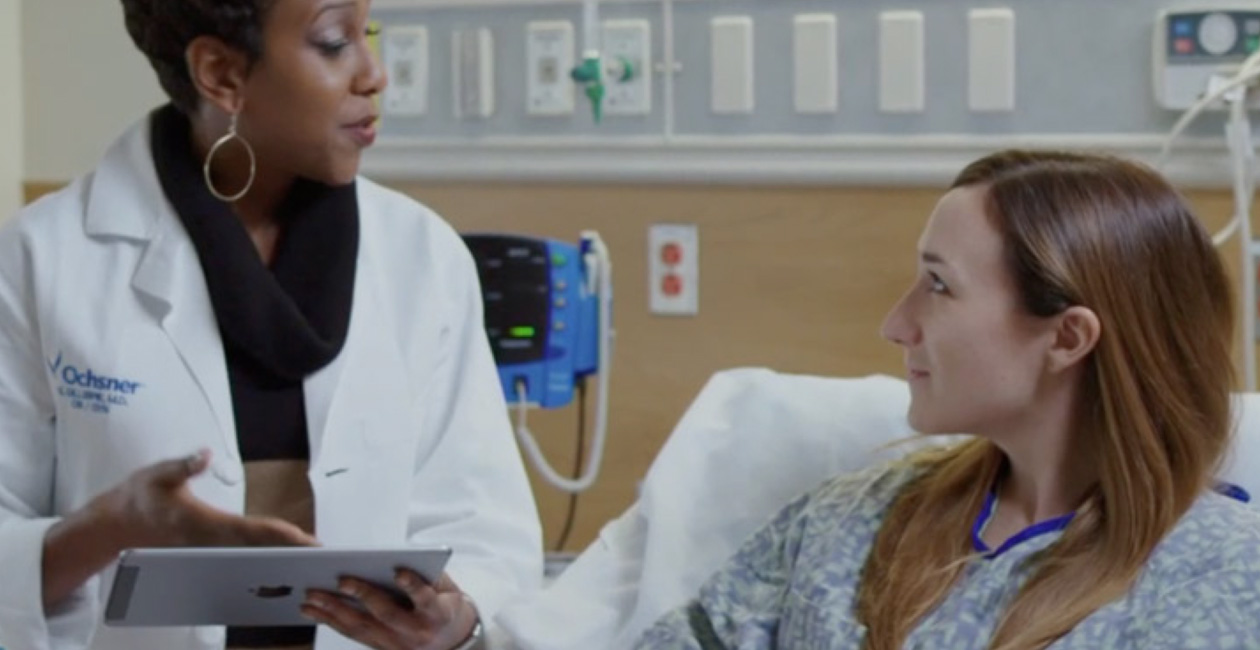 Một nhân viên y tế cầm iPad đang nói chuyện với bệnh nhân trên giường bệnh.