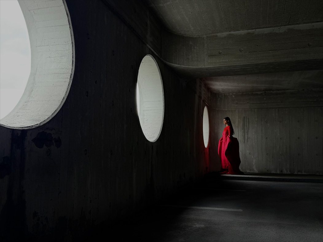 A woman wearing a long red dress stands in an empty room being photographed. The Photo app toggles through the following focal lengths to demonstrate the capabilities of the pro camera system: 200 mm, 100 mm, 48 mm, 35 mm, 28 mm, 13 mm, and macro