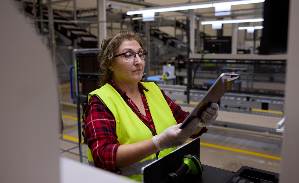An employee wearing a high-visibility vest and latex gloves inspects an iPad at a work station