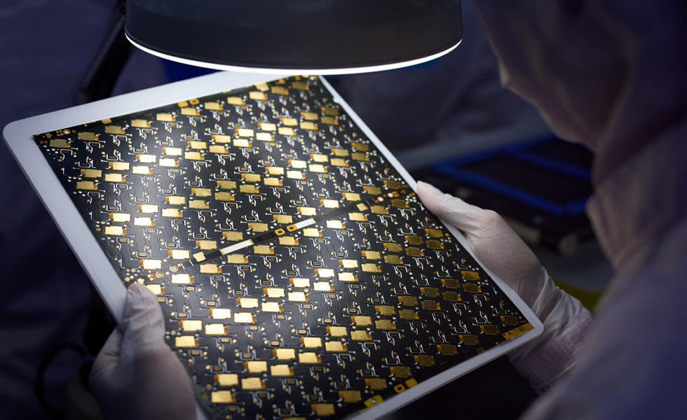 Dipendente con i guanti che ispeziona una scheda a circuito stampato usando una lampada con lente di ingrandimento