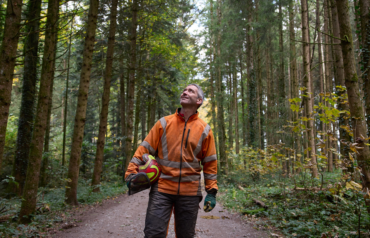 Un uomo con un giubbotto arancione ad alta visibilità e un casco di protezione in mano su un sentiero di una foresta a gestione responsabile in Svizzera