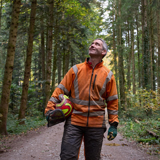 Un uomo con un giubbotto arancione ad alta visibilità e un casco di protezione in mano su un sentiero di una foresta a gestione responsabile in Svizzera