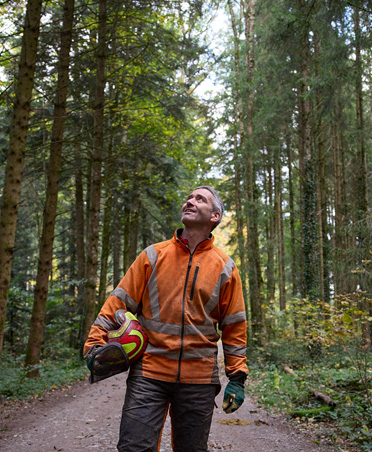 Un uomo con un giubbotto arancione ad alta visibilità e un casco di protezione in mano su un sentiero di una foresta a gestione responsabile in Svizzera
